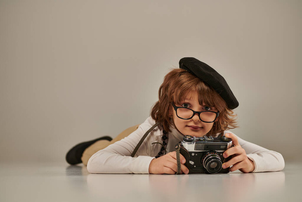 happy young photographer in beret and susenders taking photo on retro camera and lying on floor - Фото, изображение