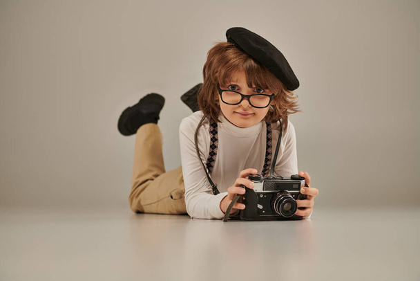 młody fotograf w berecie i okulary robiące zdjęcia przed kamerą i leżące na podłodze, kreatywny chłopiec - Zdjęcie, obraz