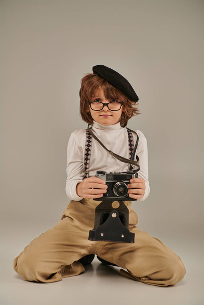 młody fotograf w berecie i okularach trzymający kamerę i siedzący na podłodze, chłopiec w szelkach - Zdjęcie, obraz
