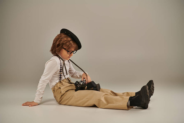 młody fotograf w berecie i okulary trzyma aparat fotograficzny i siedzi na podłodze, ładny chłopiec w szelkach - Zdjęcie, obraz