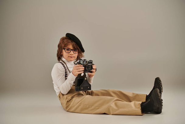 mladý fotograf v baretu a brýlích drží fotoaparát a sedí na podlaze, roztomilý kluk v podvazcích - Fotografie, Obrázek