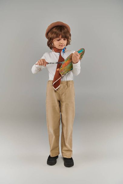 młody artysta trzyma pędzel i paletę farb, uroczy chłopiec w berecie i szalik stojący na szarym tle - Zdjęcie, obraz
