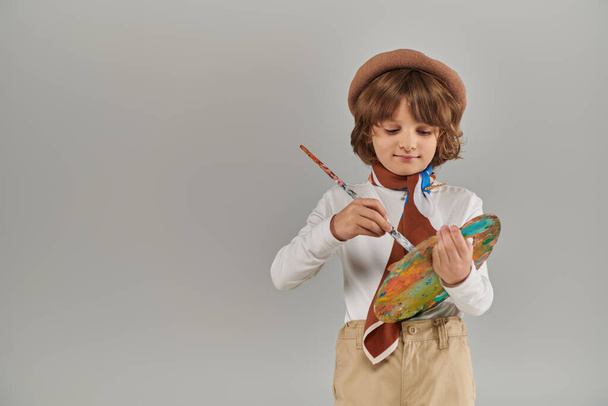 young painter holding paint brush and palette, cute kid in beret and scarf standing on grey backdrop - Fotoğraf, Görsel