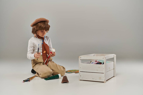 Junge kniet auf dem Boden, umgeben von Farben in Tuben und einem hölzernen Werkzeugkasten, junger Künstler in Baskenmütze - Foto, Bild
