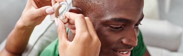 african american man smiling as his girlfriend assists with hearing aid, medical equipment banner - Photo, Image