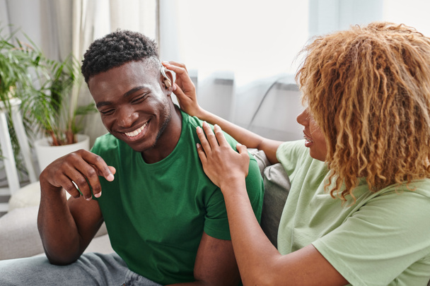 africký Američan muž se usmívá jako jeho kudrnaté přítelkyně pomáhá s naslouchátkem, lékařské vybavení - Fotografie, Obrázek