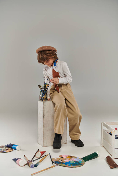 stylish artist sitting near white block with paint brushes, boy in beret and scarf in studio - Photo, image
