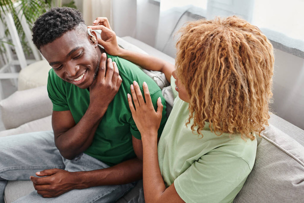 sordo afroamericano hombre sonriendo como su novia rizada ayuda con audífonos, dispositivo médico - Foto, Imagen
