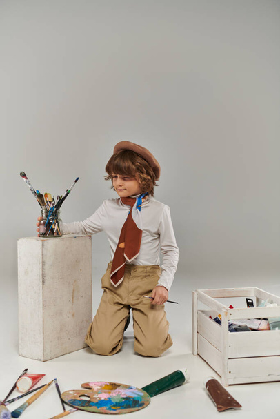 młody artysta siedzi w pobliżu białego bloku z pędzlami malarskimi, stylowy chłopiec w berecie i szalik w studio - Zdjęcie, obraz