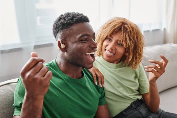 hombre afroamericano emocionado en dispositivo de audífono sentado cerca de novia feliz en aparatos ortopédicos - Foto, imagen