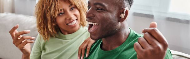 hombre afroamericano emocionado en dispositivo de audífono sentado cerca de novia feliz en frenillos, pancarta - Foto, imagen