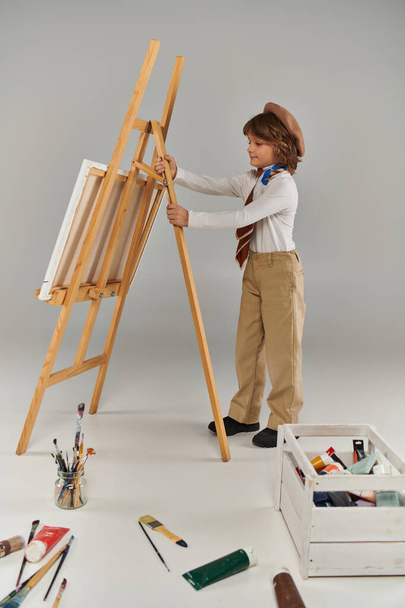 young artist adjusting his easel, surrounded by his tools and colorful paint, boy in beret and scarf - Photo, Image