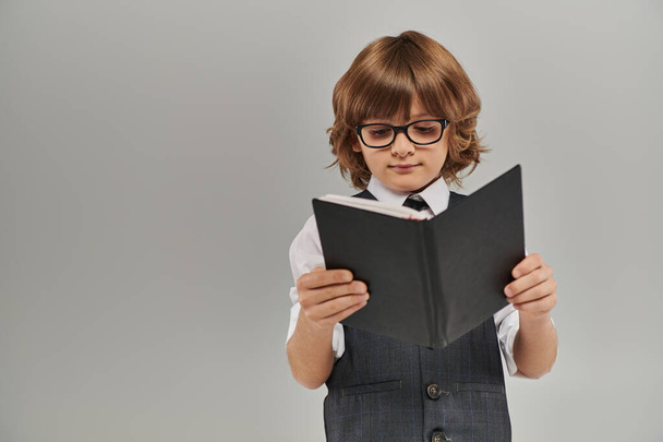 περίεργο αγόρι με γυαλιά βυθίζεται σε έναν κόσμο φαντασίας καθώς διαβάζει ένα βιβλίο για το γκρι φόντο - Φωτογραφία, εικόνα