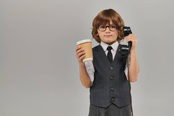 elegante jongen in bril en formele kleding houden retro telefoon en drinken in papieren beker op grijs - Foto, afbeelding