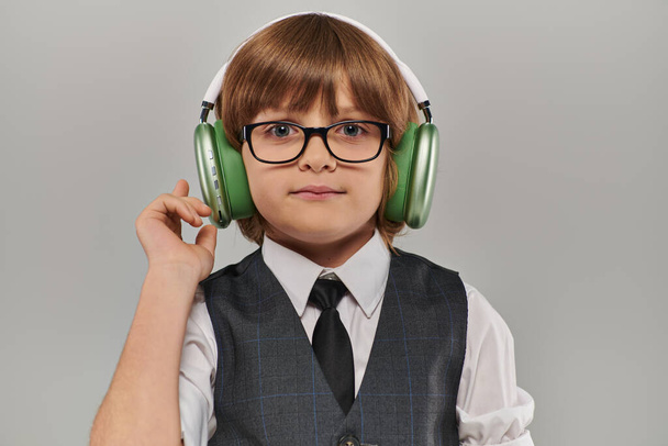 menino elegante em óculos e traje elegante com colete ouvindo música através de fones de ouvido verdes - Foto, Imagem