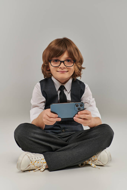 ragazzo sorridente in occhiali ed elegante abbigliamento gioco mobile su grigio, tenendo smartphone - Foto, immagini