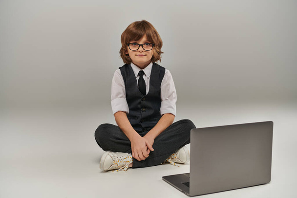 niño en traje elegante con chaleco y pantalones sentados cerca de la computadora portátil en gris, futuro hombre de negocios - Foto, imagen