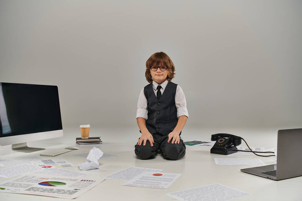 chłopiec w okularach otoczony papierami i materiałami biurowymi na szarym tle, przyszły biznesmen - Zdjęcie, obraz