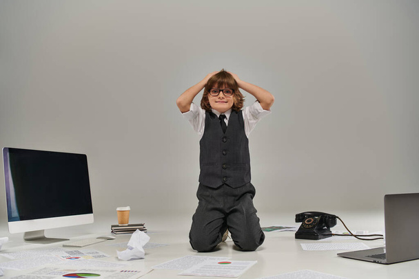 niño estresado en gafas rodeado de papeles y suministros de oficina en gris, futuro hombre de negocios - Foto, imagen