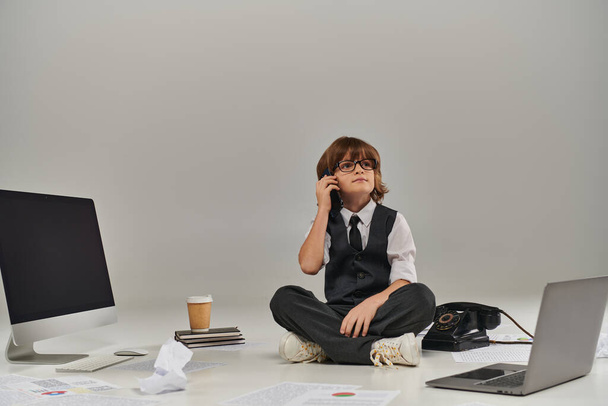 chlapec v brýlích a formální oblečení mluví na smartphone a sedí obklopen kancelářskou technikou - Fotografie, Obrázek