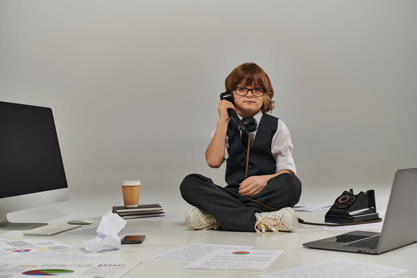 dzieciak w okularach i formalnym ubraniu rozmawiający przez telefon retro i siedzący otoczony sprzętem biurowym - Zdjęcie, obraz