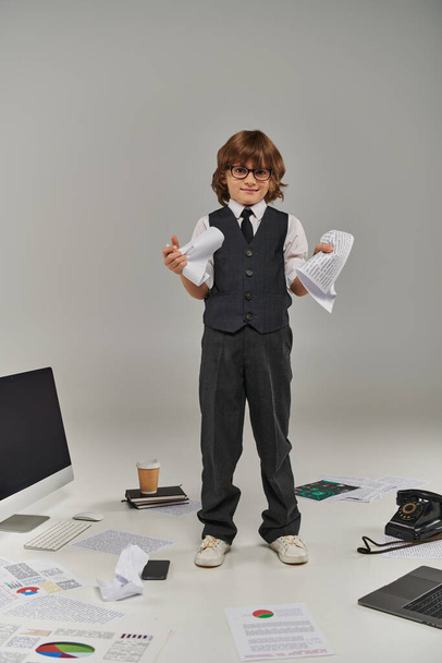 χαριτωμένο αγόρι σε γυαλιά και επίσημη φθορά που περιβάλλεται από εξοπλισμό γραφείου και συσκευές στέκεται με χαρτιά - Φωτογραφία, εικόνα
