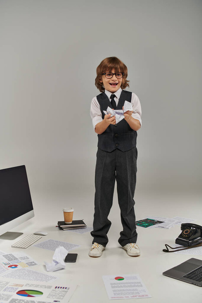 excited boy in glasses and formal wear surrounded by office equipment and devices holding papers - 写真・画像