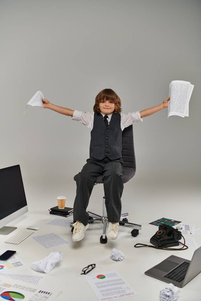Ofis malzemeleriyle çevrili, elinde kağıtlarla sandalyede oturan mutlu çocuk. - Fotoğraf, Görsel