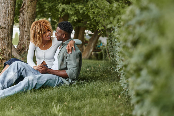 happy african american woman in casual wear hugging boyfriend while sitting together on grass - Фото, изображение
