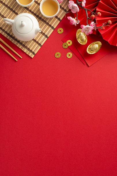 Opulent tavolo Capodanno cinese: Overhead colpo verticale cattura ventilatore tradizionale, set da tè, bacchette, tappetino di bambù, monete fortunate, buste e sakura sullo sfondo rosso, lasciando spazio per testo o annunci - Foto, immagini