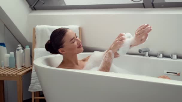 4K video görüntüsü, köpüklü banyoda neşeli bir kadın ellerini lifle yavaşça ovuyor. Huzurlu bir ortamda rahatlama ve kişisel bakım anı - Video, Çekim