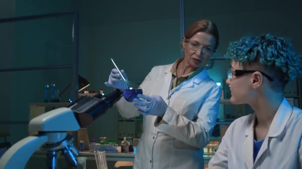 Inyección media de dos investigadoras en batas de laboratorio y gafas revisando y discutiendo muestras de soluciones azules en frasco y frasco, mientras realizan experimentos científicos - Imágenes, Vídeo