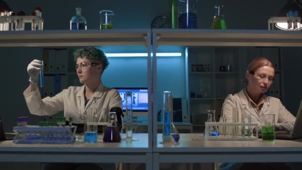 Średnie ujęcie młodej techniki żeńskiej z protezą ręki mieszającej niebieski roztwór chemiczny w fiolce, a następnie podającej go dojrzałemu kaukaskiemu naukowcowi, siedzącemu przy biurku pisząc na laptopie, podczas wykonywania badań - Materiał filmowy, wideo