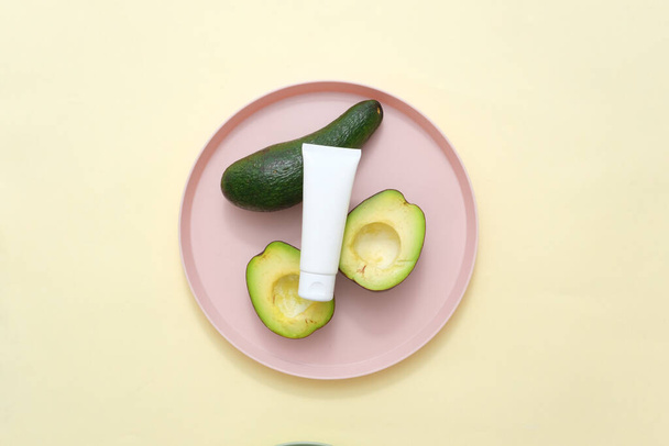 Advertising photo, mockup cosmetic tube displayed on pink ceramic dish with fresh avocados on beige background. Top view, concept for advertising organic cosmetic of avocado extract - Photo, Image