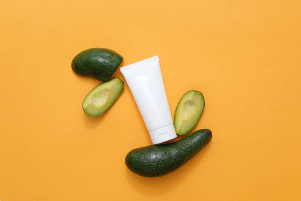 Against the yellow background, an empty plastic tube decorated with fresh slices of avocado slices. Mockup packaging, cosmetics design. Minimalist concept for advertising with organic ingredient - Photo, Image