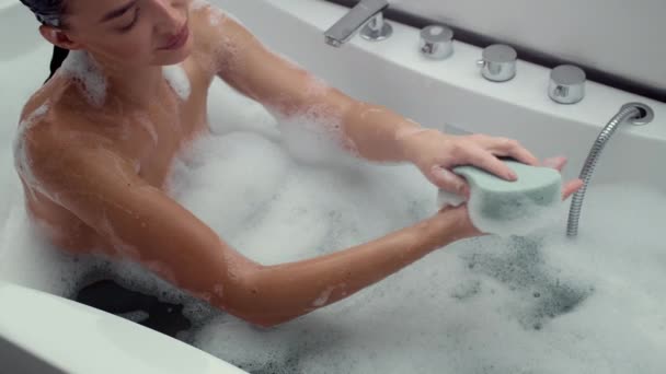 4K video beelden vrolijke vrouw in schuim gevulde badkamer schrobben haar handen met een loofah, slow motion. De scène omvat een moment van ontspanning en persoonlijke verzorging in een rustige omgeving, - Video