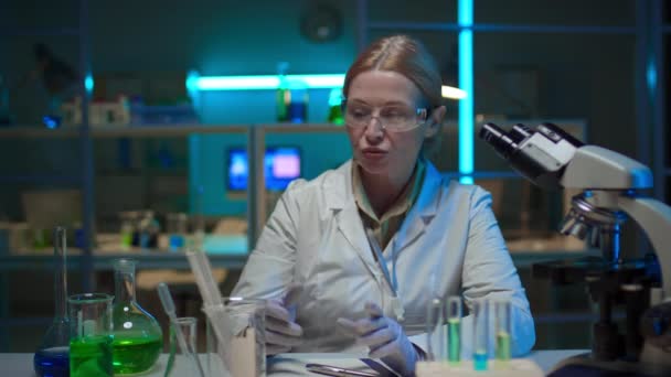 Střední záběr učitelky bělošky středního věku sedící za stolem v laboratoři, vysvětlující nové koncepty, ukazující baňku s nazelenalou chemickou tekutinou, při nahrávání online přednášky - Záběry, video