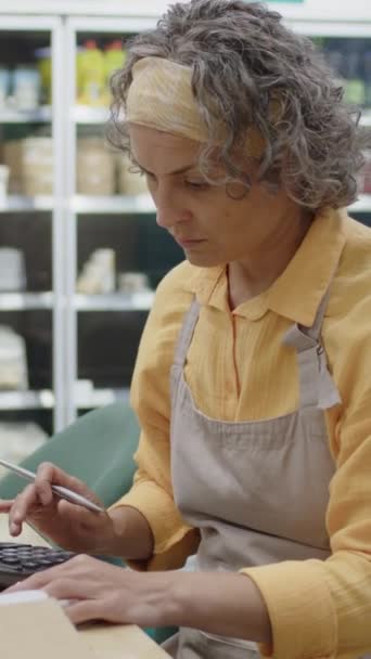 Pionowa talia w górę ujęcia białej kobiety pracującej w supermarkecie przeglądającej paragony i liczącej na kalkulator podczas regularnego audytu rano - Materiał filmowy, wideo