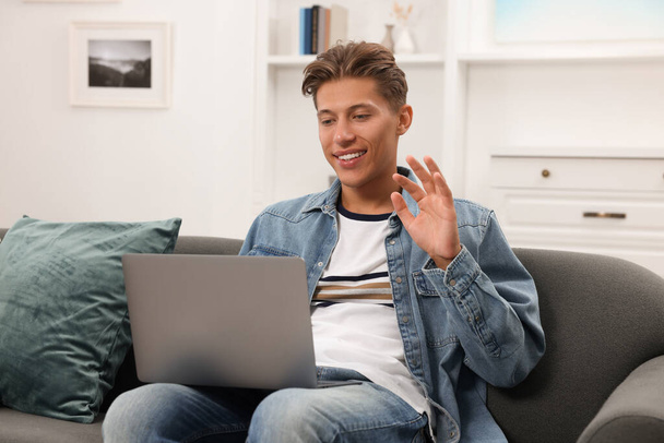 Glücklicher junger Mann mit Videochat per Laptop auf Sofa drinnen - Foto, Bild