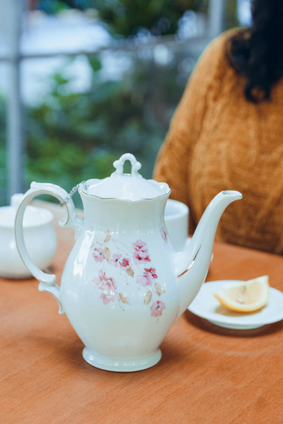 pionowy obraz czasu herbaty z nierozpoznawalną kobietą w tle i biały czajniczek porcelany z filiżanką i cytryną slajdów na stole. - Zdjęcie, obraz