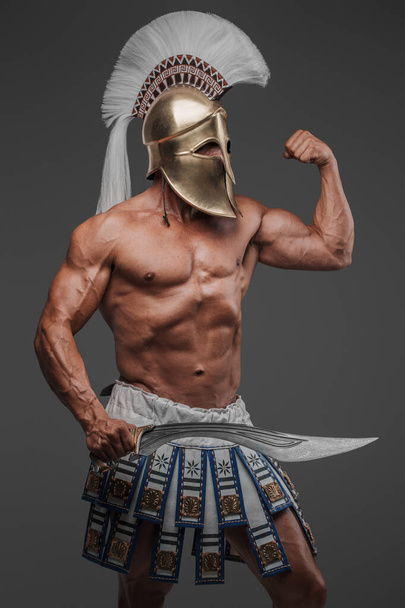 Портрет древнегреческого гоплита, позирующего в студии с обнаженным мускулистым туловищем, в шлеме и держащего гладиуса, установленного на сером фоне - Фото, изображение