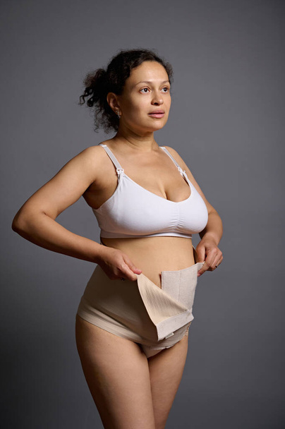 Auténtico retrato de estudio de una mujer multiétnica, madre joven que se pone un vendaje elástico después de una cesárea, mirando hacia otro lado, aislado sobre un fondo de estudio gris. Concepto de cuidado de la piel y el cuerpo - Foto, imagen