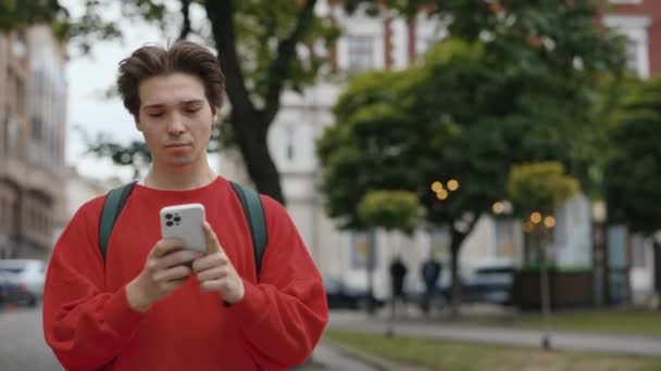 Beau jeune homme caucasien en vêtements décontractés utilisant Smartphone tout en marchant sur la rue City. Le gars dehors. Textos avec des amis. Attrayant Male Scrolling Apps. Concept technologique - Séquence, vidéo
