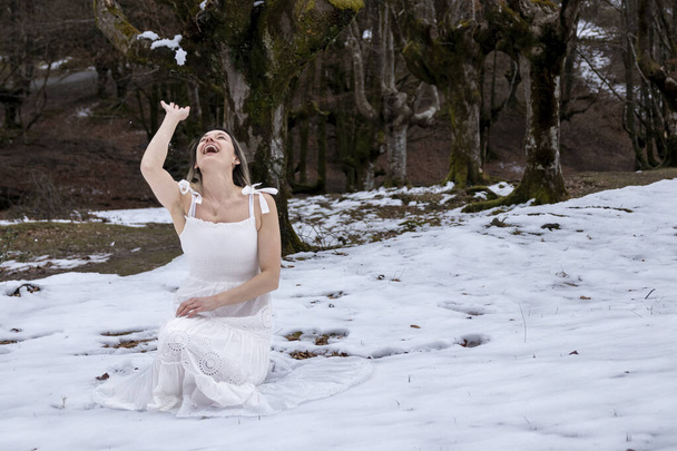 woman in a white dress is laughing joyfully while sitting in a snowy forest, creating a serene and peaceful scene - Foto, Imagem