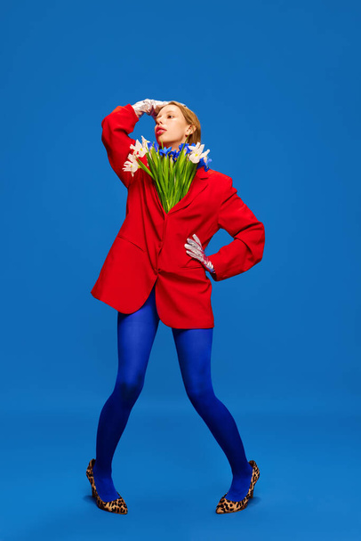 Λαμπερή νεαρή γυναίκα με μακιγιάζ. Παράξενο κορίτσι με λουλούδια σε ασυνήθιστο, παράξενο, κόκκινο σακάκι που ποζάρει σε φόντο στούντιο μπλε χρώματος. Έννοια της υψηλής μόδας, στυλ και αίγλη, ομορφιά, διαφήμιση - Φωτογραφία, εικόνα