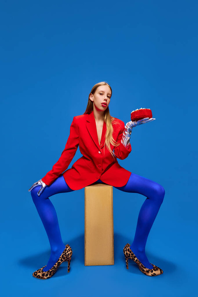 Fashion standards. Young lady posing in unusual, freaky, bright outfit, wearing blue tights, red jacket and animal printed heels holding birthday cake. Concept of high fashion, style, glamour, beauty. - Photo, Image