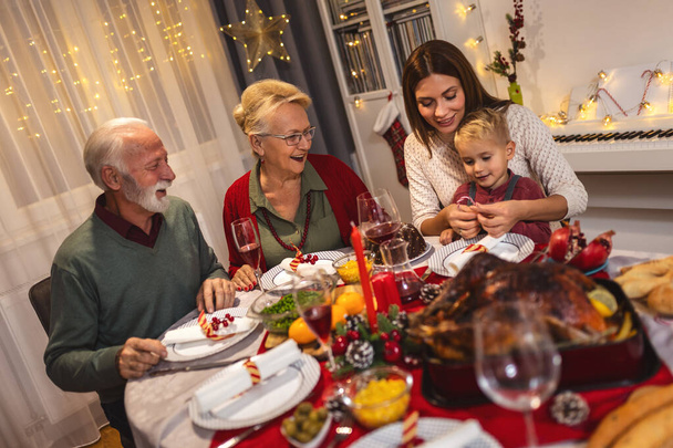 Mutlu bir aile Noel 'de büyükannesi ve büyükbabasıyla yemek yiyor. Ev yapımı yemekler yemek, içecekler içmek. Ev yeni yıl arifesi ve Noel için dekore edilmiştir. - Fotoğraf, Görsel