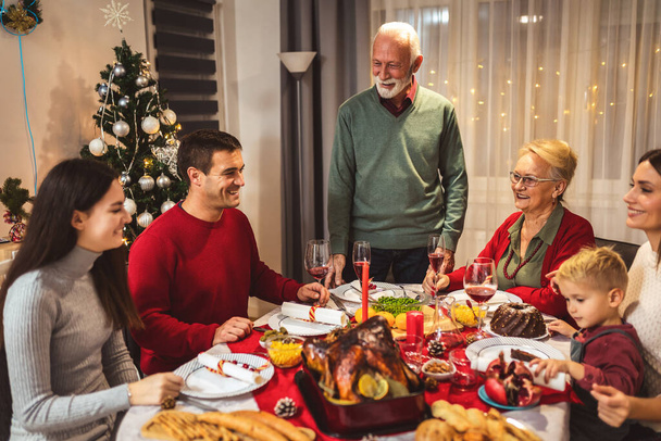 Ο παππούς κάνει πρόποση στο τραπέζι. Η οικογένεια γιορτάζει μαζί τα Χριστούγεννα τρώγοντας σπιτικό φαγητό. - Φωτογραφία, εικόνα