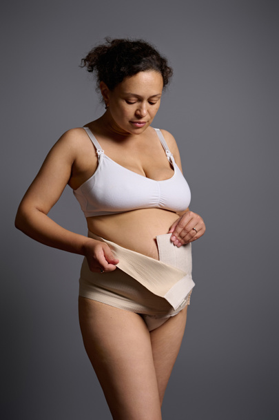 Latin Amerikalı orta yaşlı çıplak bir kadının dikey portresi, sezaryen sonrası iç çamaşırlı genç bir anne, elastik bandaj giyiyor, gri stüdyo arka planında izole edilmiş. Doğum sonrası. Hamilelik sonrası - Fotoğraf, Görsel
