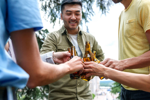 grupo de diversos amigos tintineo botellas de cerveza fuera, incluyendo feliz asiático hombre. - Foto, imagen